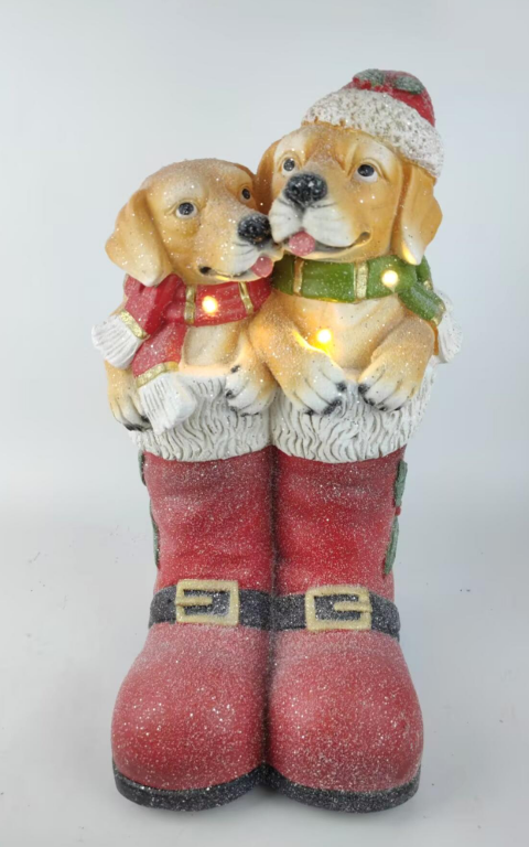 A festive Christmas figurine featuring two golden retriever dogs hugging inside a pair of red Santa boots with white fur trim and warm yellow string lights. The dogs wear red and green scarves and a red-and-white Santa hat.
