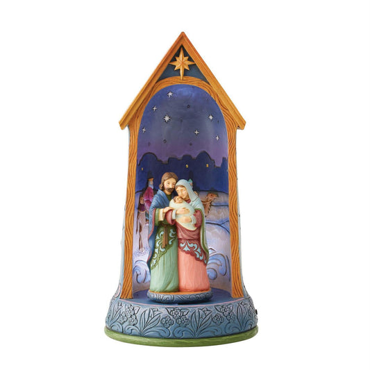 A hand-painted nativity scene figurine housed in a decorative wooden-style stable frame. The scene depicts Mary, Joseph, baby Jesus, and a shepherd under a starry night sky, with a blue patterned base.