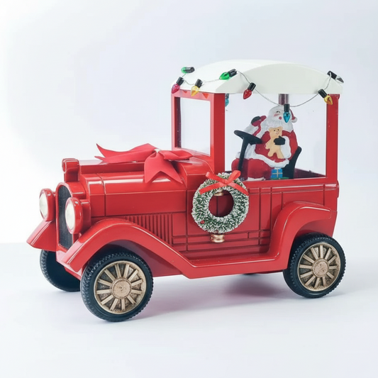 A red vintage toy car decorated for Christmas, featuring a white canopy with hanging lights, a green wreath on the side, and a Santa Claus figure driving the vehicle.
