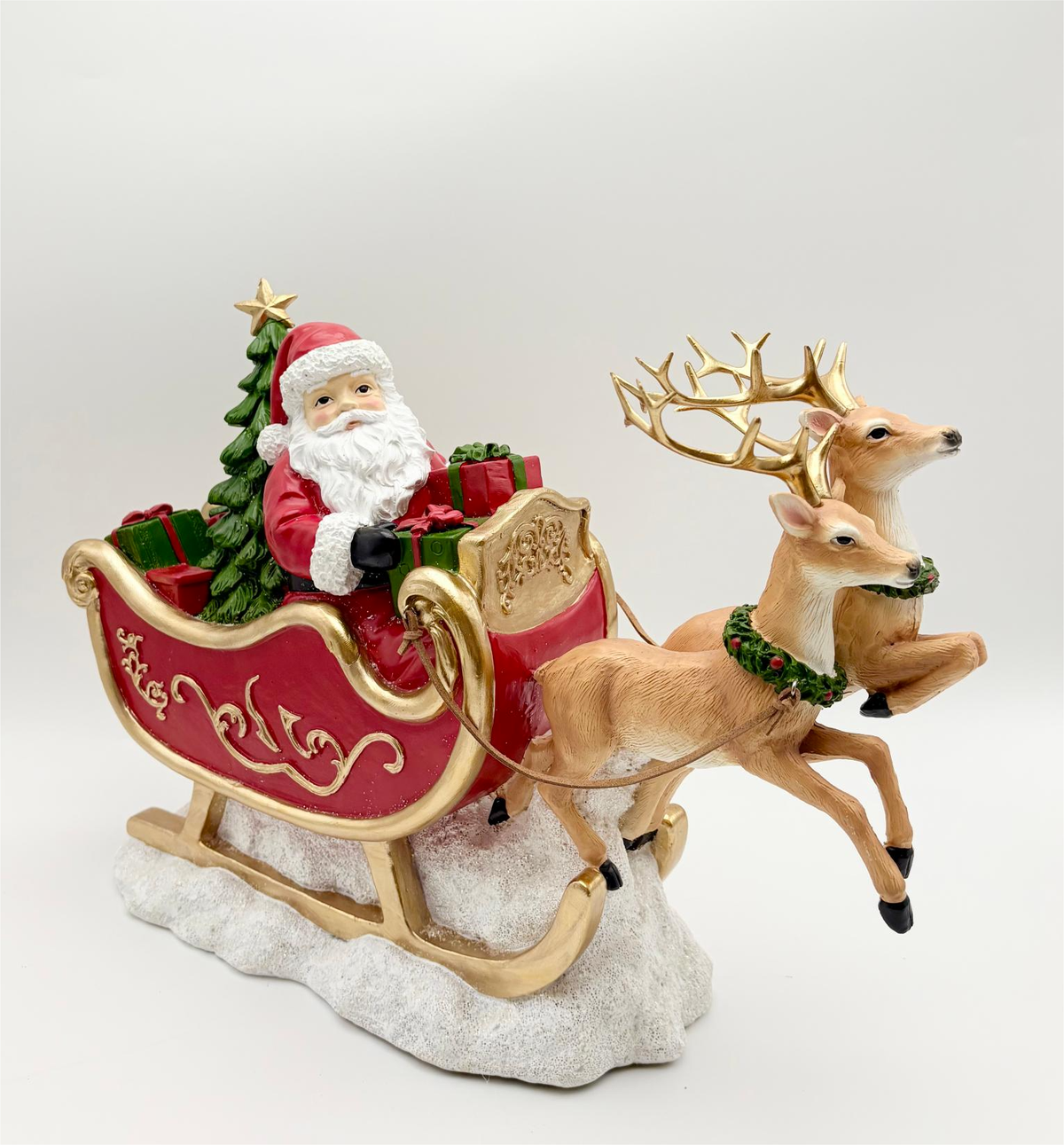 A festive Santa Claus in a red sleigh pulled by two brown reindeer, set against a plain light gray studio background. The sleigh is red with gold trim, Santa wears a red hat and white beard, and the reindeer have gold antlers with green garlands.