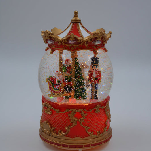 A festive Christmas-themed carousel music box snow globe. The clear glass globe is filled with glitter and miniature holiday figures, including Santa Claus and a reindeer, under a red and gold canopy.