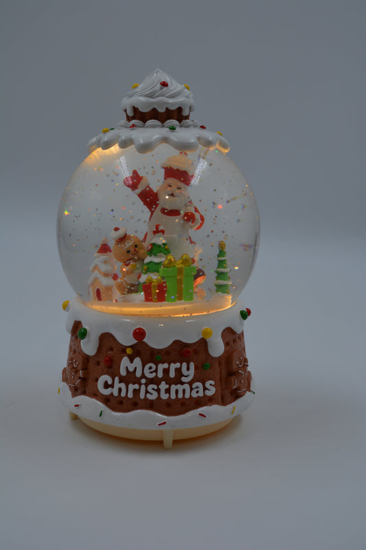 A festive Christmas snow globe figurine shaped like a gingerbread cake, featuring a Santa Claus and teddy bear scene inside, with 'Merry Christmas' written on the front, set against a neutral gray background.