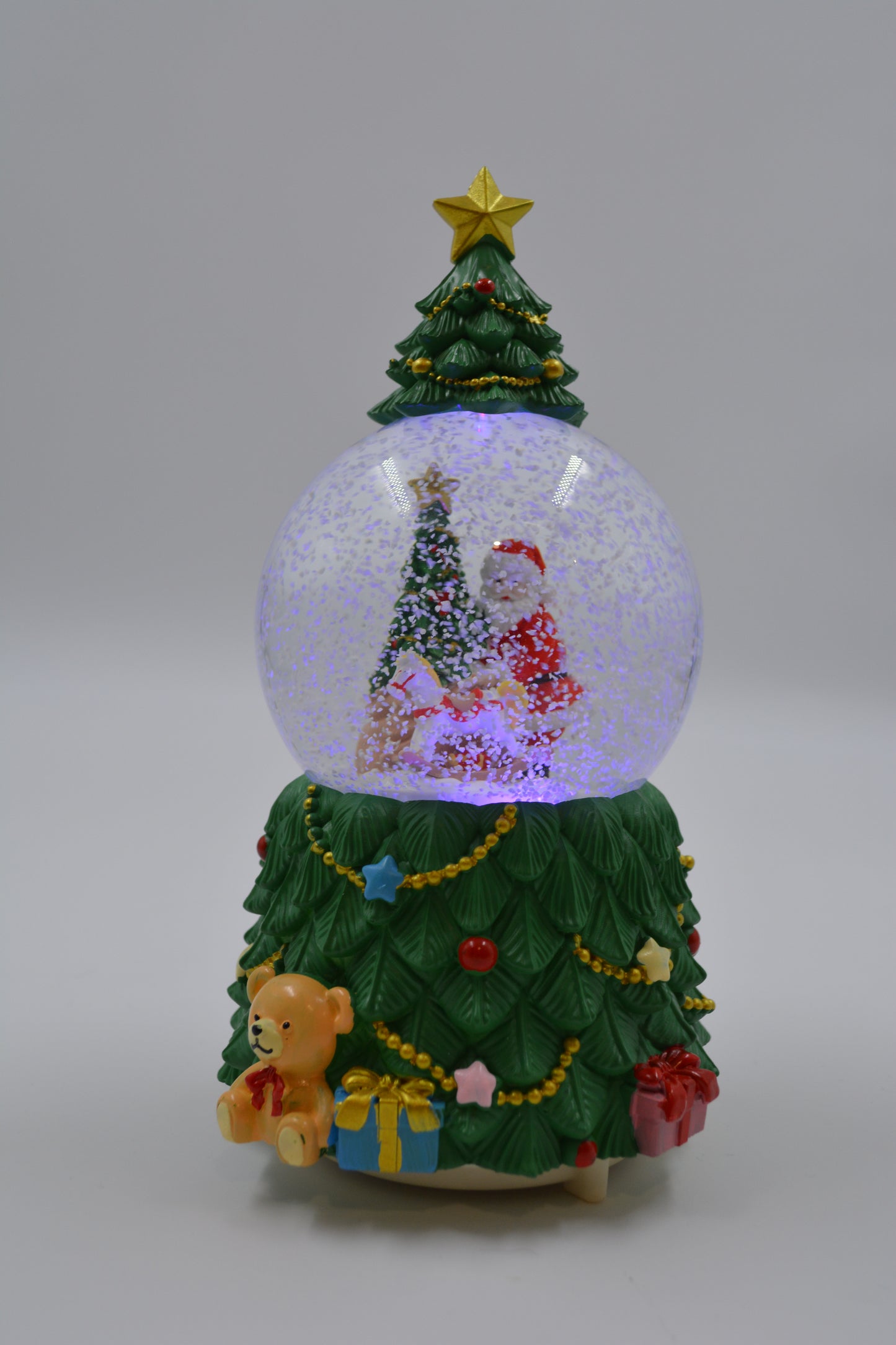 A Christmas-themed snow globe figurine with a green tree base decorated with ornaments, a teddy bear, and presents, surrounding a clear globe filled with white glitter and a miniature Santa Claus scene, shown on a plain light gray background.