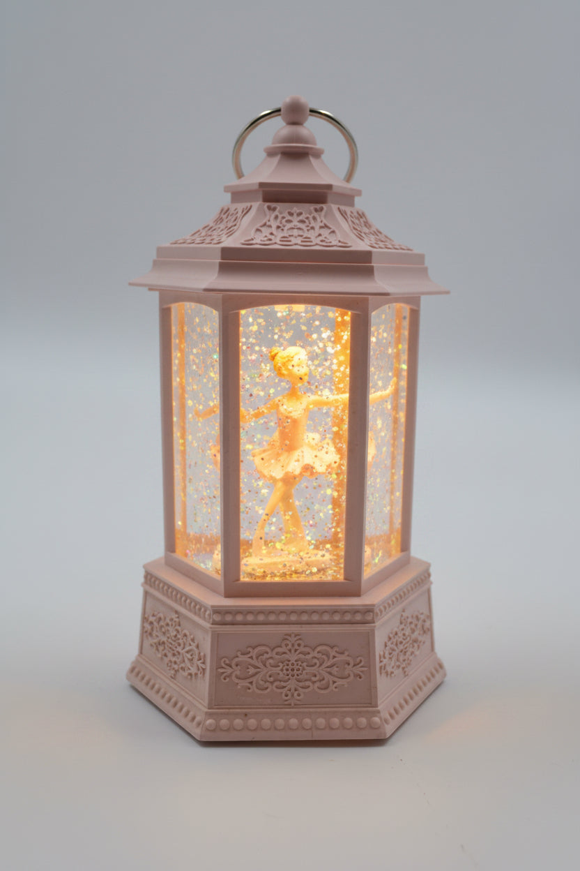 A pink decorative lantern with a ballerina figurine inside, illuminated with a warm light against a neutral gray background.