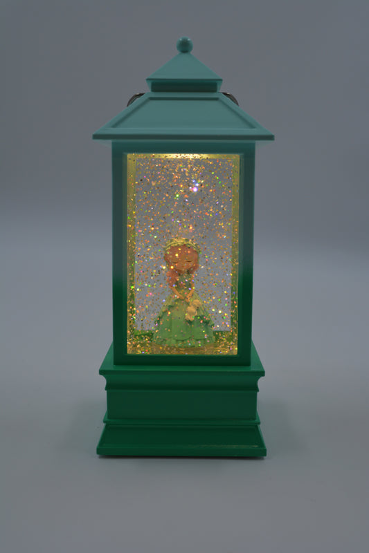 A green lantern-shaped decorative object with a glitter-filled interior and a small gold-and-green figurine inside, photographed against a smooth gray studio background.