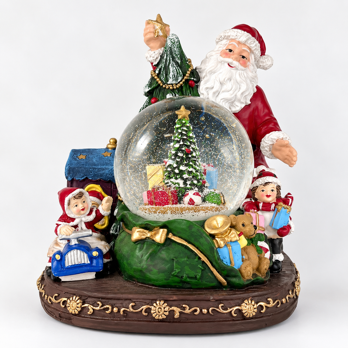 A festive Christmas snow globe figurine featuring Santa Claus, a decorated tree, gifts, a blue car, a teddy bear, and a green sack on a decorative base, photographed against a plain light background.