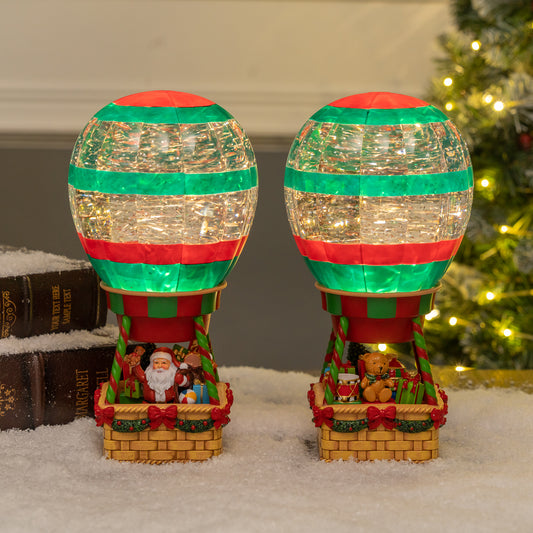 Two Christmas-themed hot air balloon lights on a snowy surface with a decorated tree in the background. Christmas snow globe