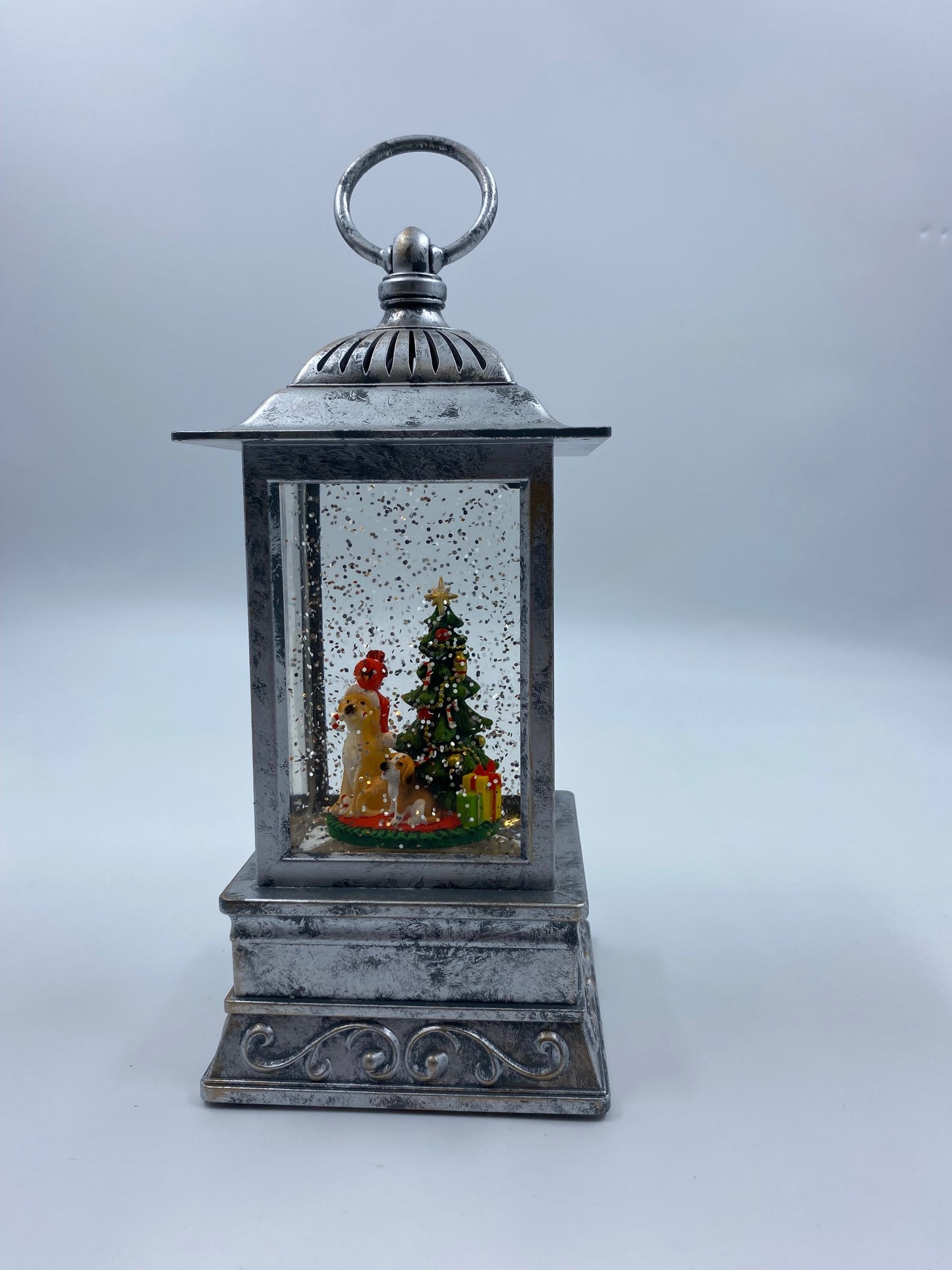 A vintage-style silver lantern snow globe decoration featuring a miniature Christmas tree, Santa Claus, and a dog inside, set against a plain light gray background.