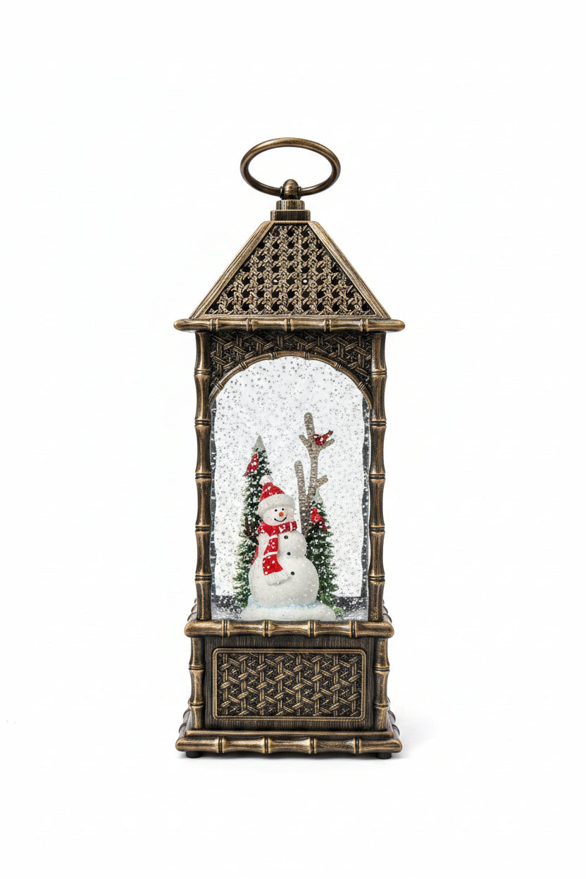 A decorative lantern-shaped snow globe with an ornate bronze-toned metal frame and a small snowman figure inside, photographed on a kitchen countertop with a white sheet of paper behind it.