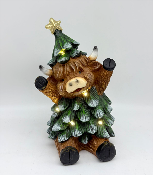 A Highland cow figurine dressed in a Christmas tree outfit with a green tree-shaped hat decorated with a gold star and small warm white lights, set against a plain light gray background.
