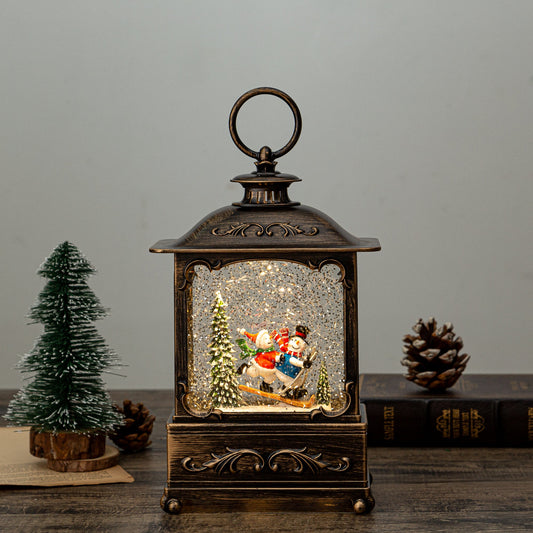 A rectangular lantern with an antique-gold/bronze frame, containing a Christmas-themed scene with a snowman, trees, and decorations, illuminated from within.