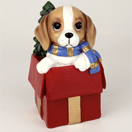A decorative figurine of a beagle puppy peeking out of a red gift box with a blue and gold striped scarf and a small green tree on its head, photographed against a plain light background.