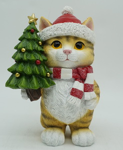 A festive ginger cat figurine wearing a red and white Santa hat and matching striped scarf, holding a small decorated Christmas tree on a plain light background.