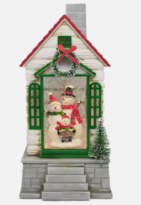 A holiday-themed lantern featuring a snow-covered cottage with Christmas decorations, including a green garland, a red bow, and snowmen in the display window.