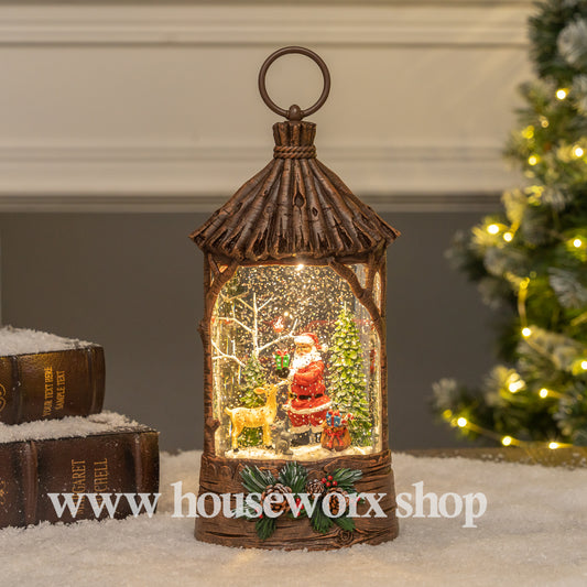Decorative lantern with a Christmas scene inside, featuring Santa Claus and a reindeer, on a snowy surface with a blurred Christmas tree in the background. Straw hut christmas snow globe