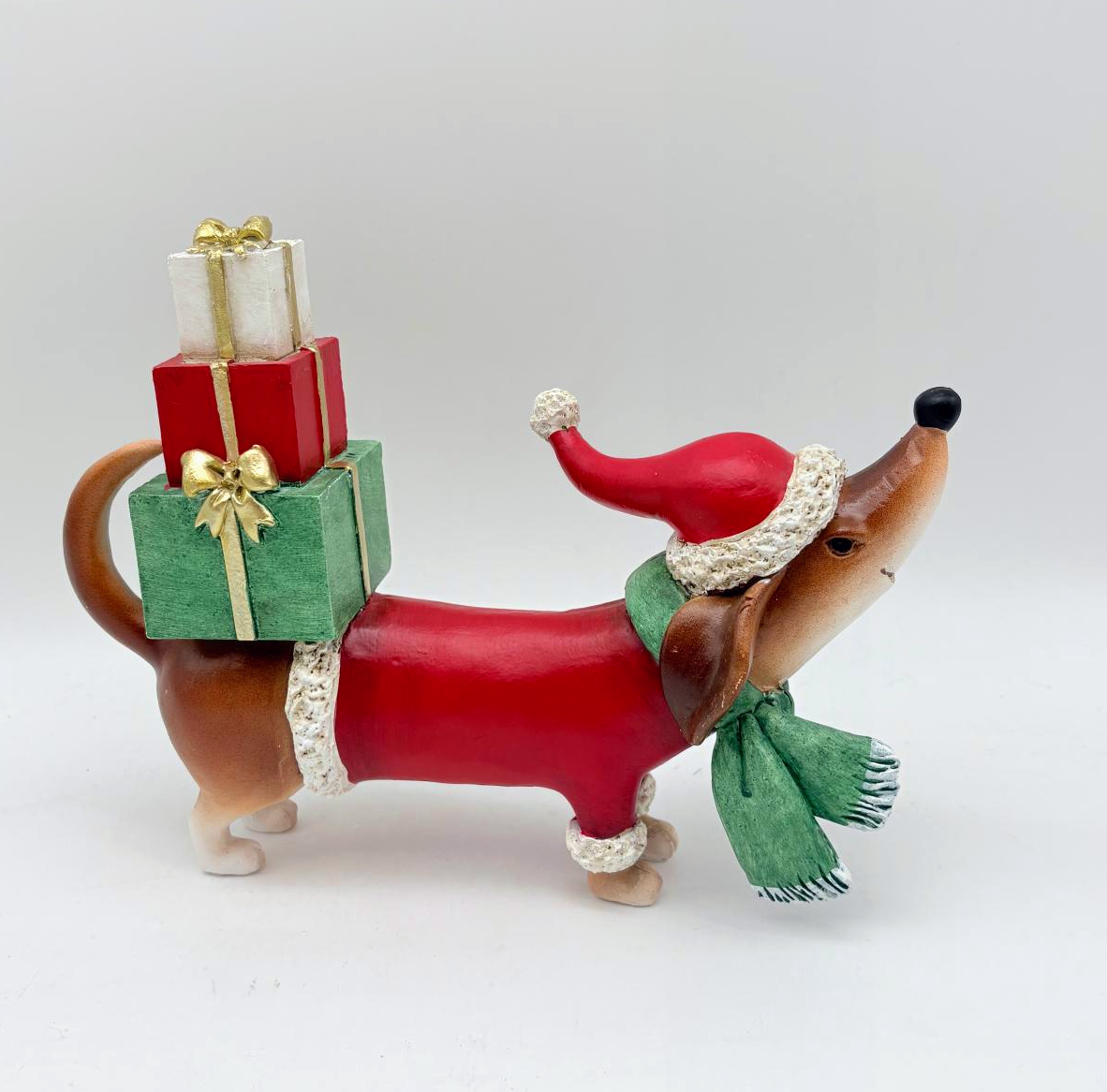 A dachshund figurine wearing a red Santa hat and green scarf, carrying a stack of three wrapped Christmas presents on its back, photographed against a plain light gray background.