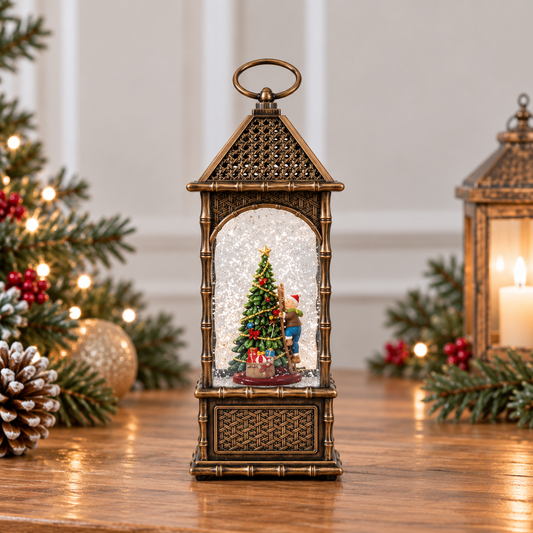 A decorative lantern-shaped Christmas ornament sits on a wooden table, surrounded by holiday greenery, pinecones, and warm lights. The lantern has an antique bronze finish and contains a miniature Christmas tree scene with a small figurine under a clear, snow-like dome.