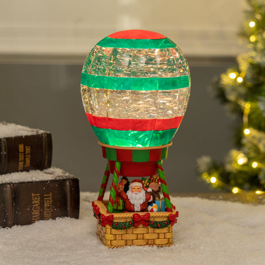 Decorative Christmas hot air balloon with lights on a surface with books and a blurred Christmas tree in the background. Christmas snow globe