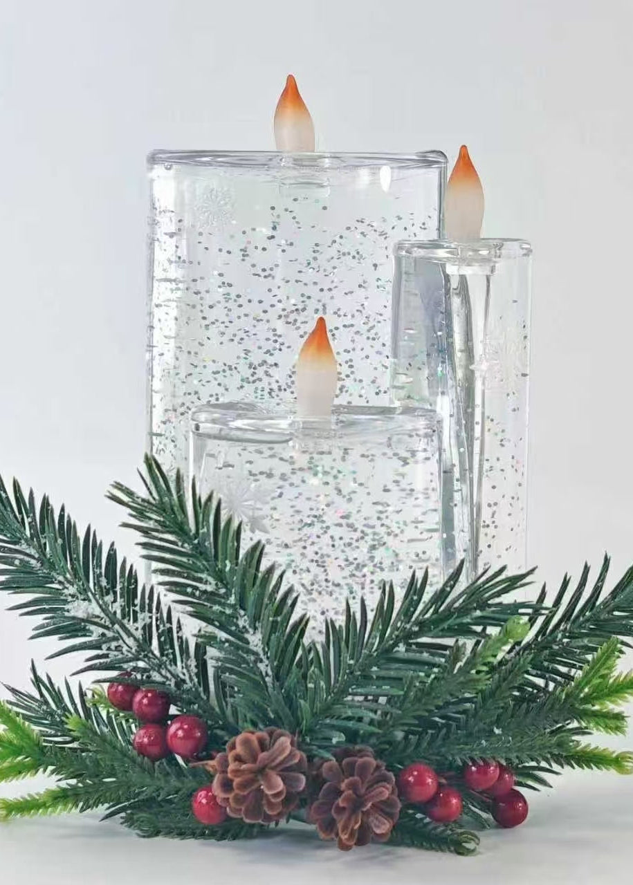 Three clear glass pillar candle holders with silver glitter and flame-shaped LED candles, styled with green pine branches, pinecones, and red berries on a white background.