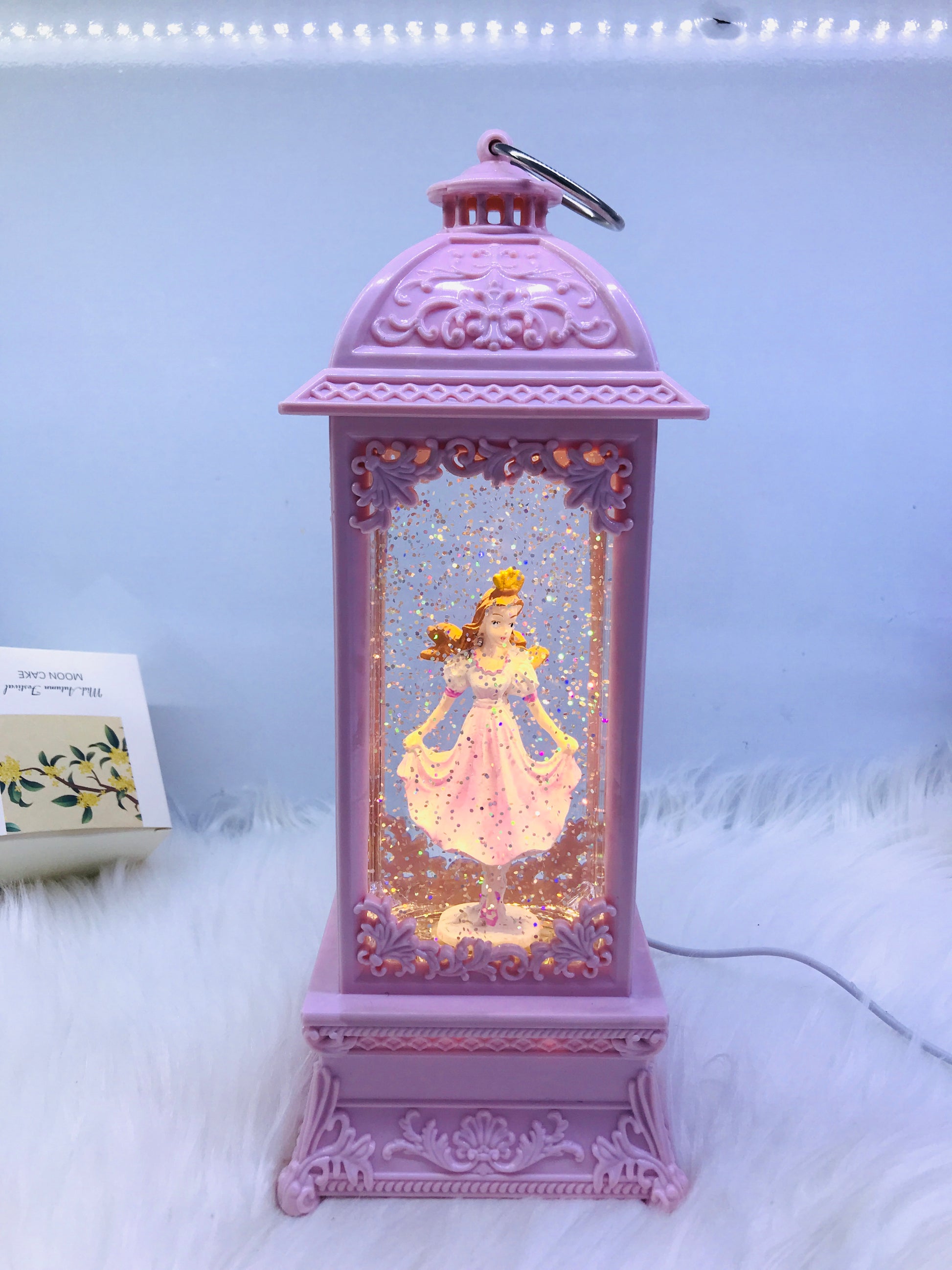 A lavender purple decorative lantern with a hanging loop, featuring a glowing fairy figure inside and a silver wire cord extending from the side, set on a white faux fur surface.