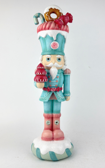 A festive nutcracker figurine dressed in a turquoise and pink holiday outfit, standing on a white and turquoise base. The top of the hat is decorated with a candy cane, donut, and red berries, and the nutcracker holds a red patterned sack.