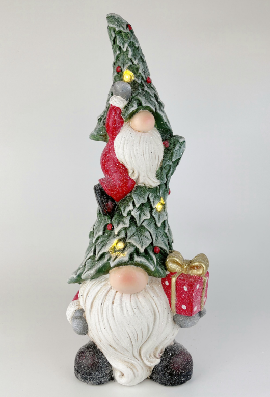 A Christmas gnome figurine with a tall, decorated tree-shaped hat, white beard, red coat, and a small red gift box with a gold bow, photographed against a plain light gray background.