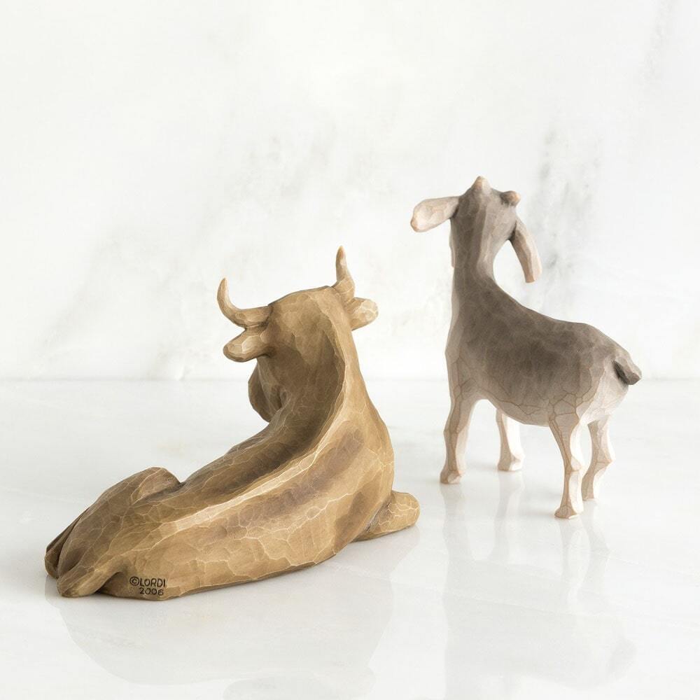 Two small decorative animal figurines on a glossy white surface against a soft white marble-like background. The larger figurine in the foreground is tan and brown with horns, and the smaller figurine behind it is light gray with pale legs.