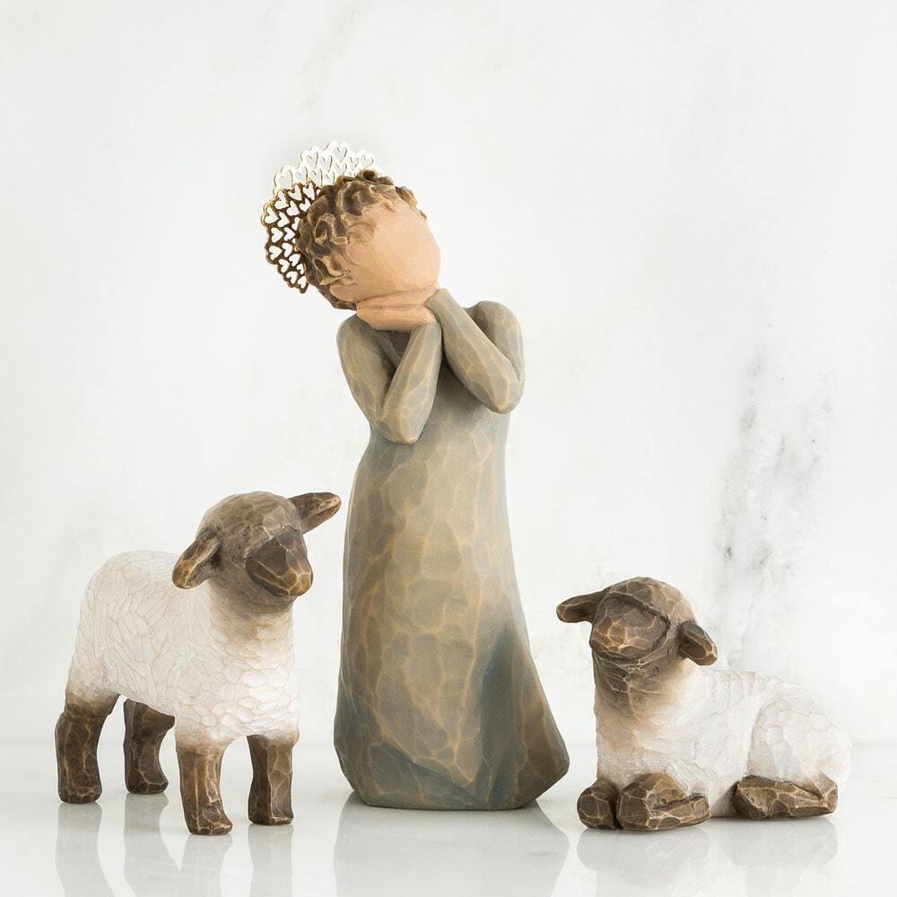 A set of three Willow Tree figurines on a reflective white surface against a light gray marble background: a standing woman in a long robe with her head tilted back, wearing a tiara, flanked by two sheep, one standing and one lying down.