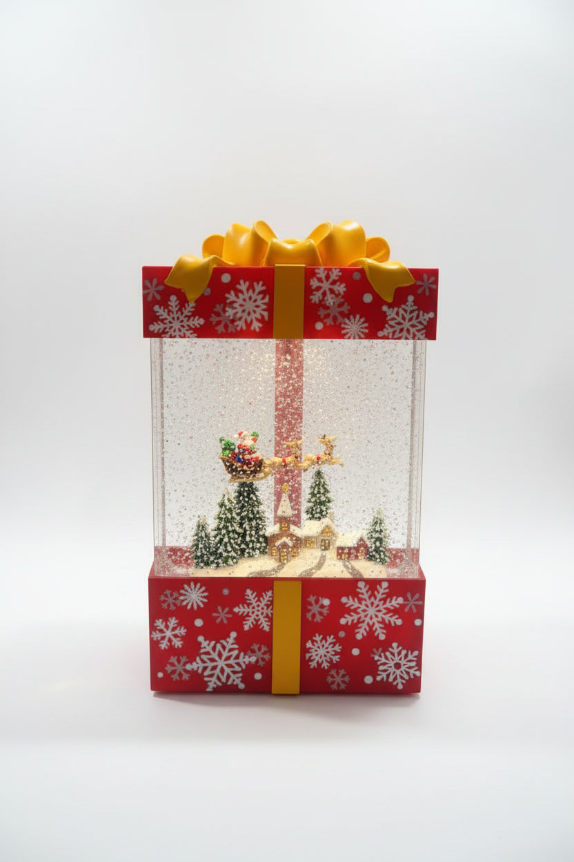 A Christmas-themed gift box snow globe decoration with a red and white patterned box, yellow ribbon, and glitter-filled clear globe containing miniature holiday trees, a golden angel, and Santa.
