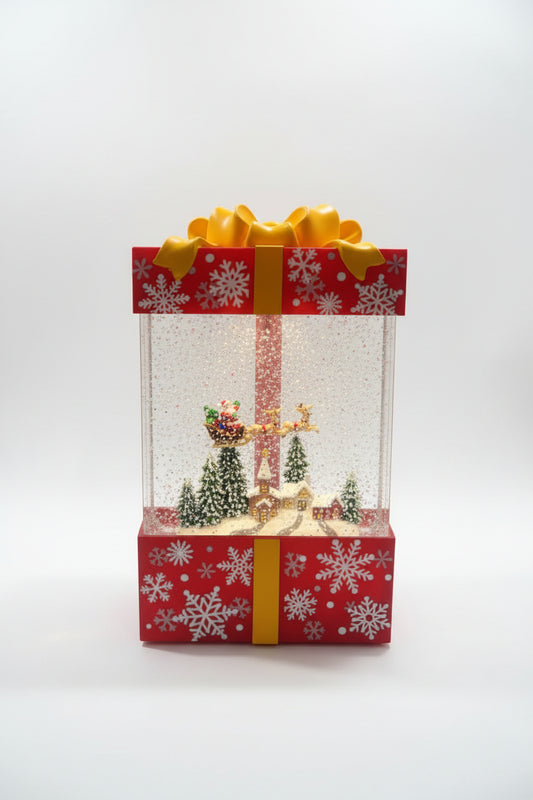 A Christmas-themed gift box snow globe decoration with a red and white patterned box, yellow ribbon, and glitter-filled clear globe containing miniature holiday trees, a golden angel, and Santa.