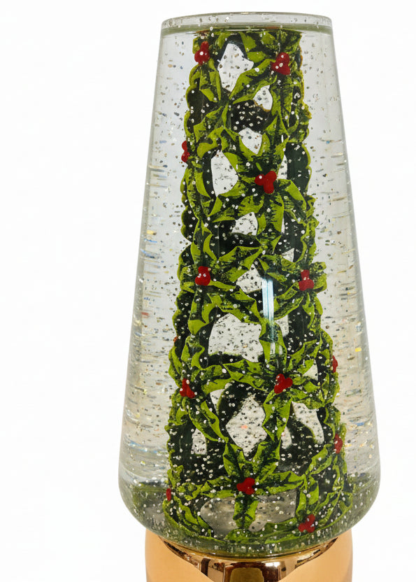 A close-up of a clear glass decorative container or vase with a green holly and red berries pattern inside, filled with small gold glitter-like specks, sitting on a dark countertop.