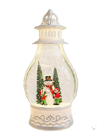 A decorative snow globe lantern with a white ornate base and lid, featuring a miniature snowman and children scene inside with small green trees, shown on a plain white background.