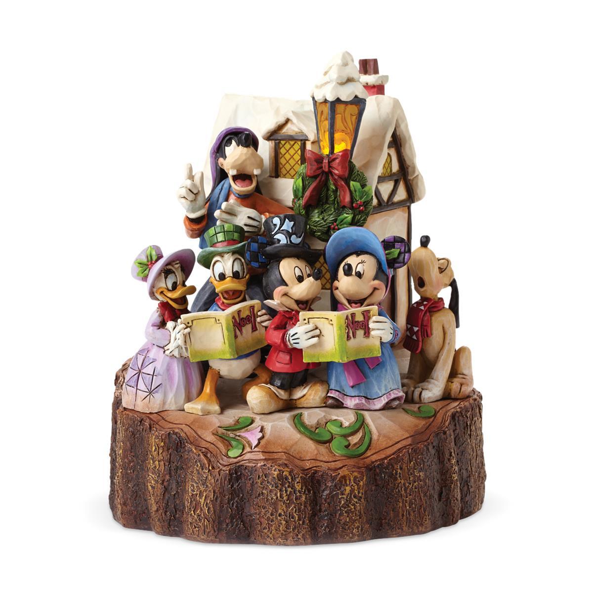 A Disney Christmas-themed figurine featuring Mickey Mouse, Minnie Mouse, Donald Duck, Goofy, Pluto, and Daisy Duck gathered around a small house on a tree-slice base, with a lantern and holiday wreath detail, photographed on a white background.