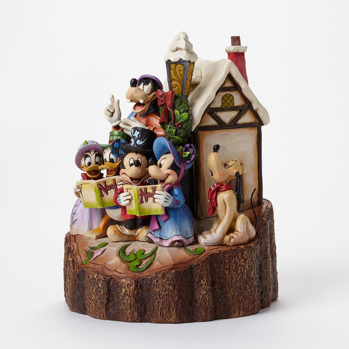 Disney Jim Shore collectible figurine of Mickey, Minnie, Goofy, Donald, Daisy, Pluto, and Donald Duck gathered around a snowy house, holding an 'M' tree ornament sign against a white background.