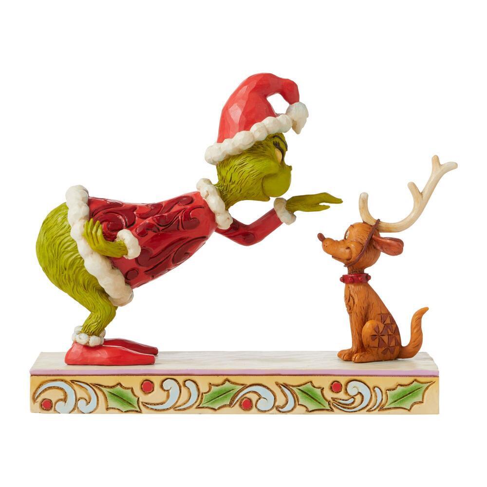 A Christmas figurine featuring the Grinch in a red Santa outfit leaning toward a brown dog with deer antlers, mounted on a decorated holiday base against a plain white background.