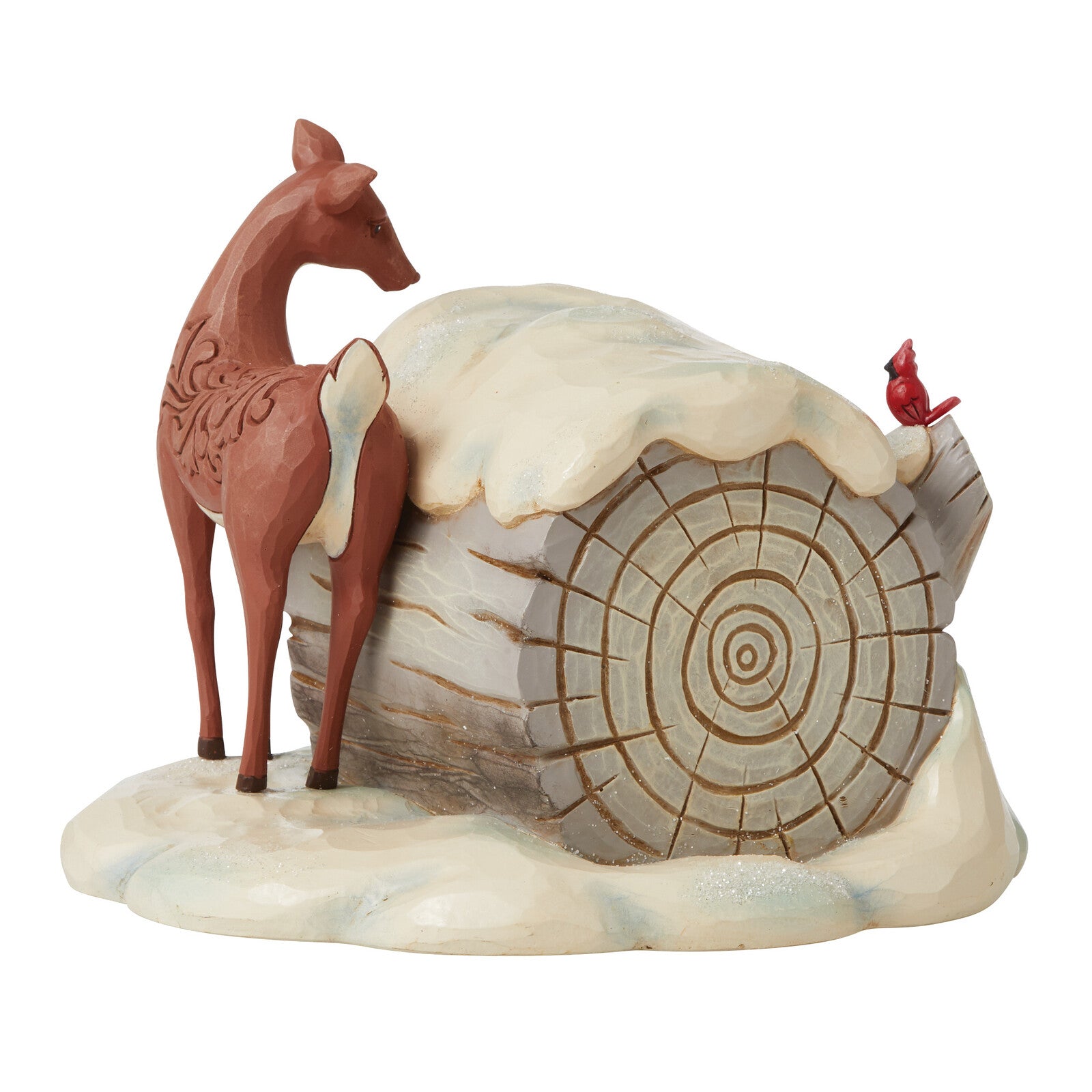 A decorative winter figurine featuring a reddish-brown deer standing beside a log covered with white faux snow, with a small red bird perched on the log, all set on a matching snowy base against a plain white background.