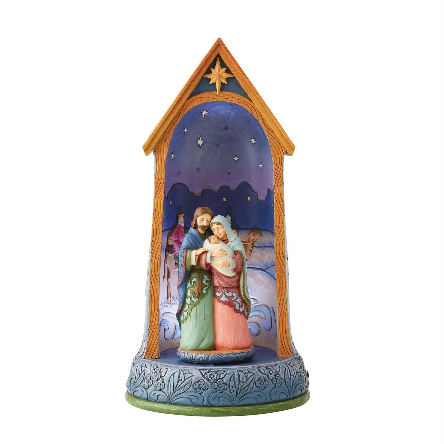 A hand-painted nativity scene figurine housed in a decorative wooden-style stable frame. The scene depicts Mary, Joseph, baby Jesus, and a shepherd under a starry night sky, with a blue patterned base.