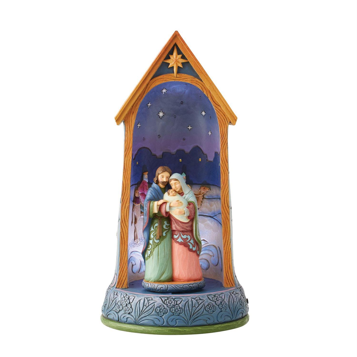 A hand-painted nativity scene figurine housed in a decorative wooden-style stable frame. The scene depicts Mary, Joseph, baby Jesus, and a shepherd under a starry night sky, with a blue patterned base.