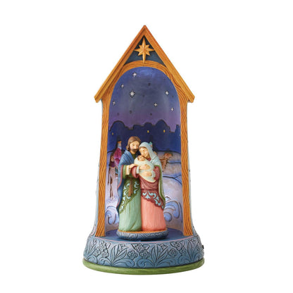 A hand-painted nativity scene figurine housed in a decorative wooden-style stable frame. The scene depicts Mary, Joseph, baby Jesus, and a shepherd under a starry night sky, with a blue patterned base.