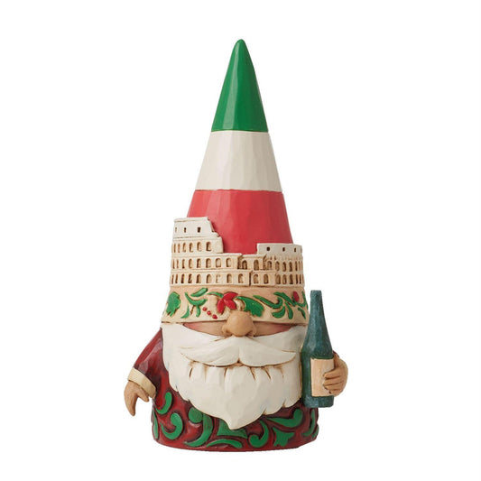 A decorative gnome figurine with a tall pointed hat in green, white, and red horizontal stripes, holding a dark green bottle. The gnome has a white beard, a red and green patterned coat, and a beige brick-patterned hat band with green holly details, shown on a plain white background.
