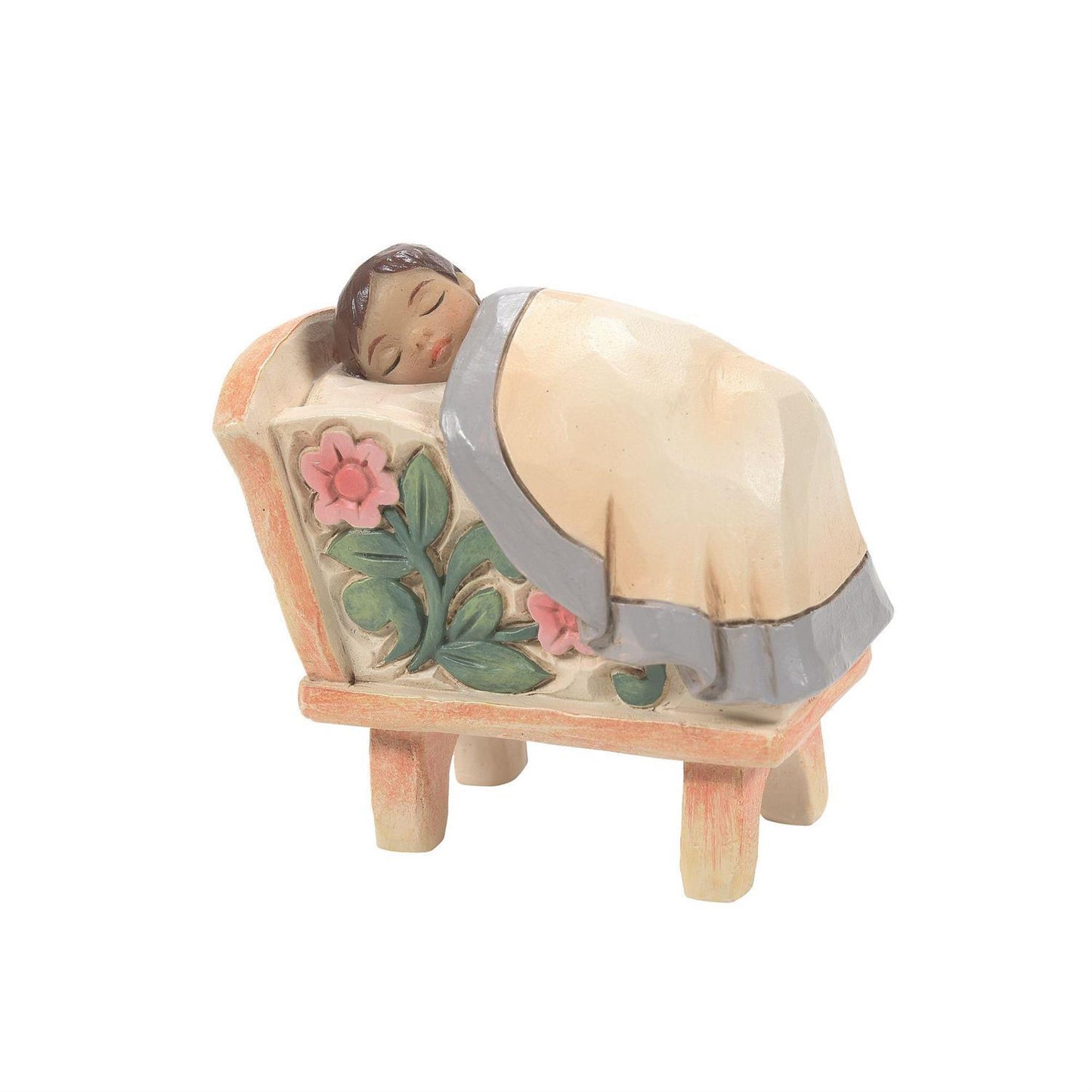 A hand-painted wooden figurine of a sleeping baby in a cradle, featuring a cream-colored blanket with pink trim, green leaves, and pink flowers on the side, set against a plain white background.