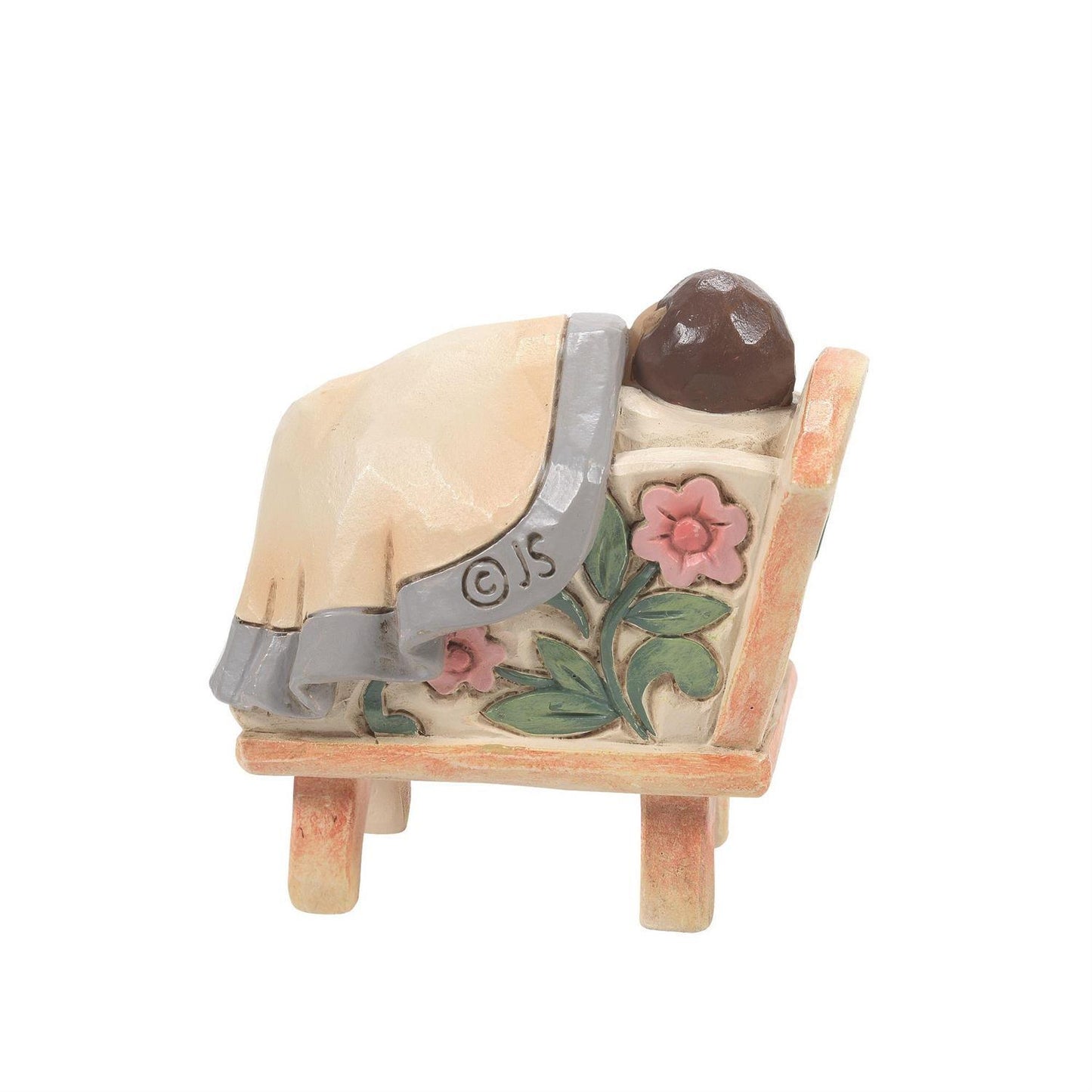 A small decorative figurine of a person lying down under a cream-colored blanket with pink flowers and green leaves, set on a small bed-like base against a plain white background.