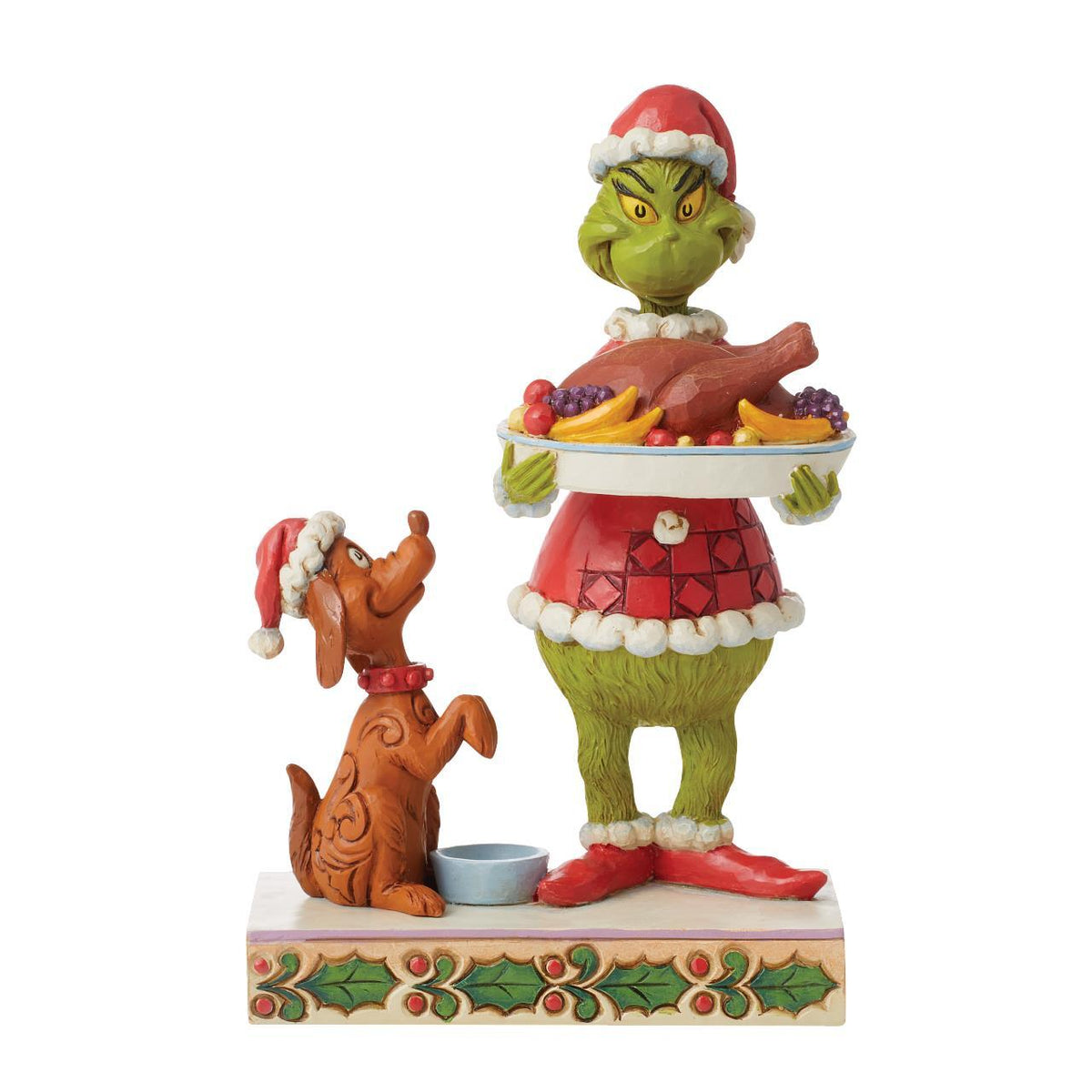 A Grinch and Santa's Dog holiday figurine displayed on a wooden table in front of a decorated Christmas tree with warm bokeh lights. The Grinch stands on one foot holding a tray with roasted turkey and fruit, while Santa's Dog sits beside him on a decorated base with holly accents.
