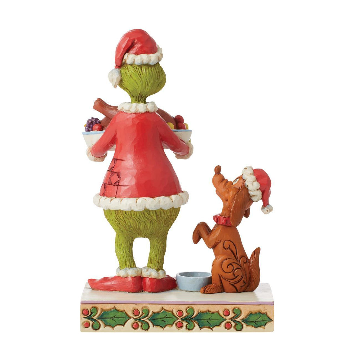 A Grinch and Santa's Dog holiday figurine displayed on a wooden table in front of a decorated Christmas tree with warm bokeh lights. The Grinch stands on one foot holding a tray with roasted turkey and fruit, while Santa's Dog sits beside him on a decorated base with holly accents.
