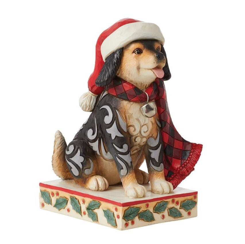 A decorative figurine of a dog wearing a red and white Santa hat, a red plaid scarf, and a black-and-white patterned coat, sitting on a holly-patterned base against a plain white background.
