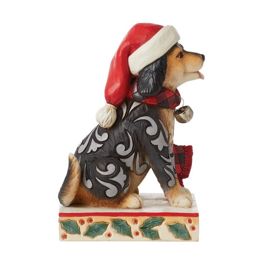 A decorative figurine of a dog wearing a red Santa hat, a red-and-black plaid scarf, and a black-and-white patterned coat, standing on a holly-accented base against a plain white background.