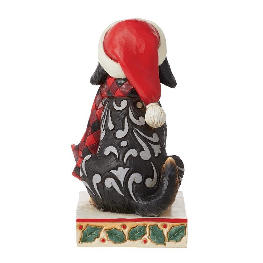 A Christmas-themed figurine of a black dog sitting on a decorated base, wearing a red Santa hat and a red plaid scarf, photographed against a plain white background.