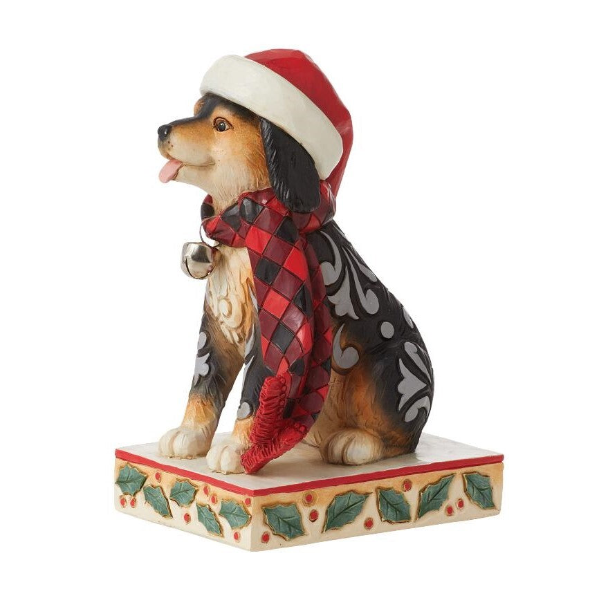 A decorative figurine of a dog wearing a red and white Santa hat and a red-and-black plaid scarf with gray patterned sleeves, sitting on a holly-accented base against a plain white background.