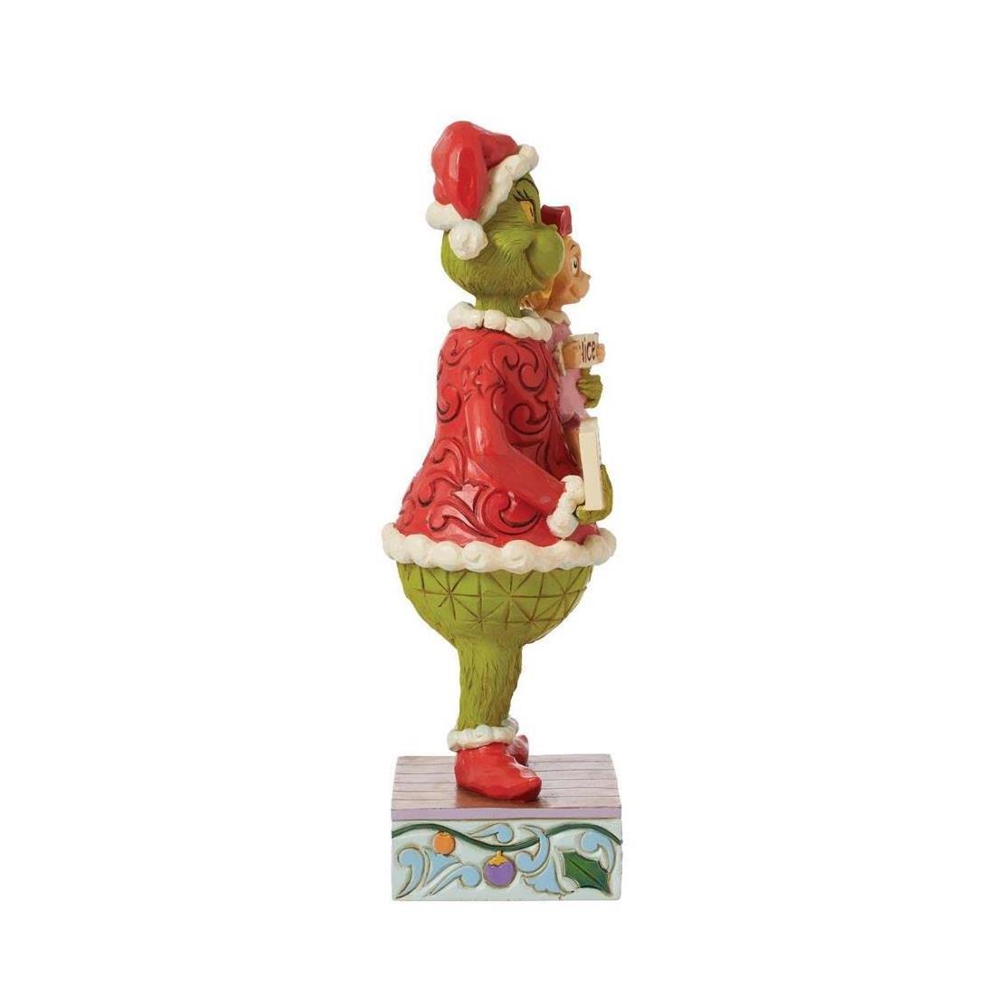 A Grinch figurine wearing a red Santa hat and red Santa coat, standing on a decorated gift box against a plain white background.