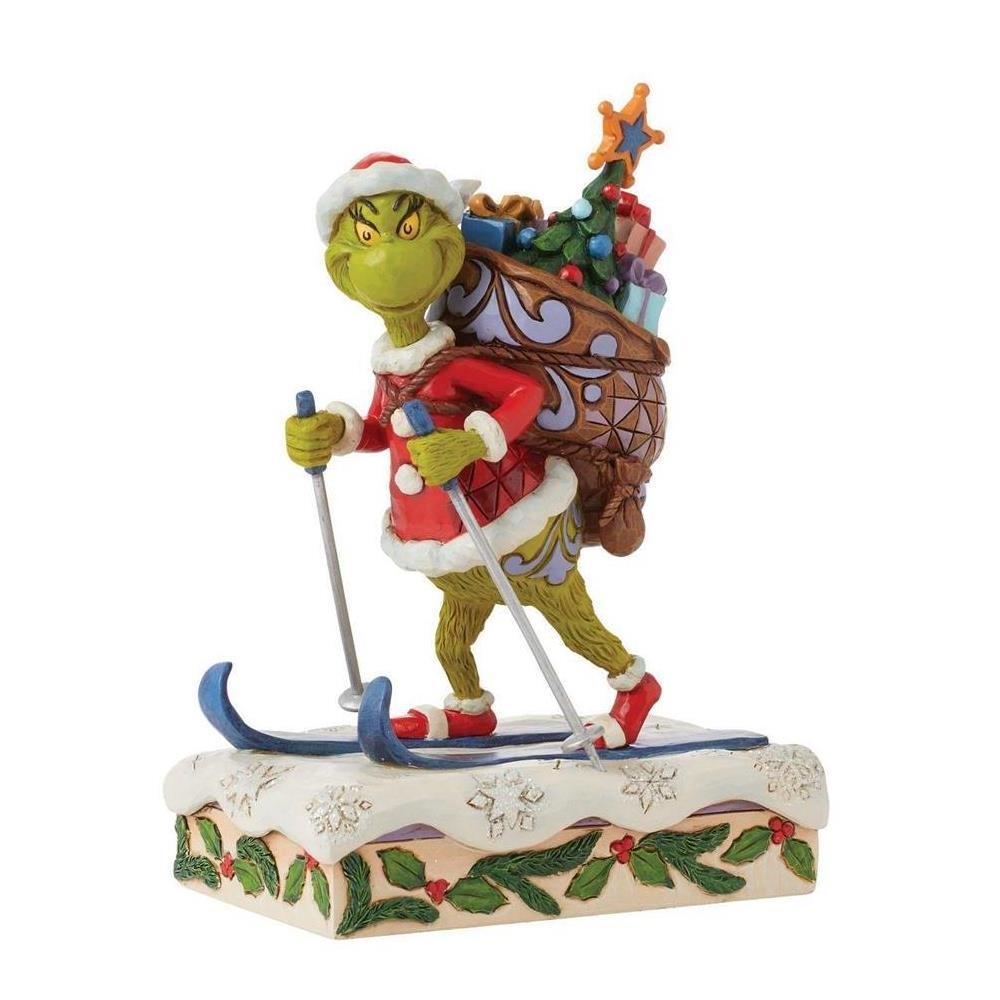 A Grinch figurine depicted skiing with poles and a large Christmas sack filled with presents and a tree, set on a snow-covered base with holly and berries, photographed on a plain white background.